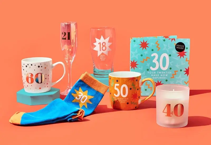 Collection of colorful birthday-themed items against an orange background, including numbered mugs, glasses, socks, a candle, and a gift box labeled '30 Your twenties have now expired!